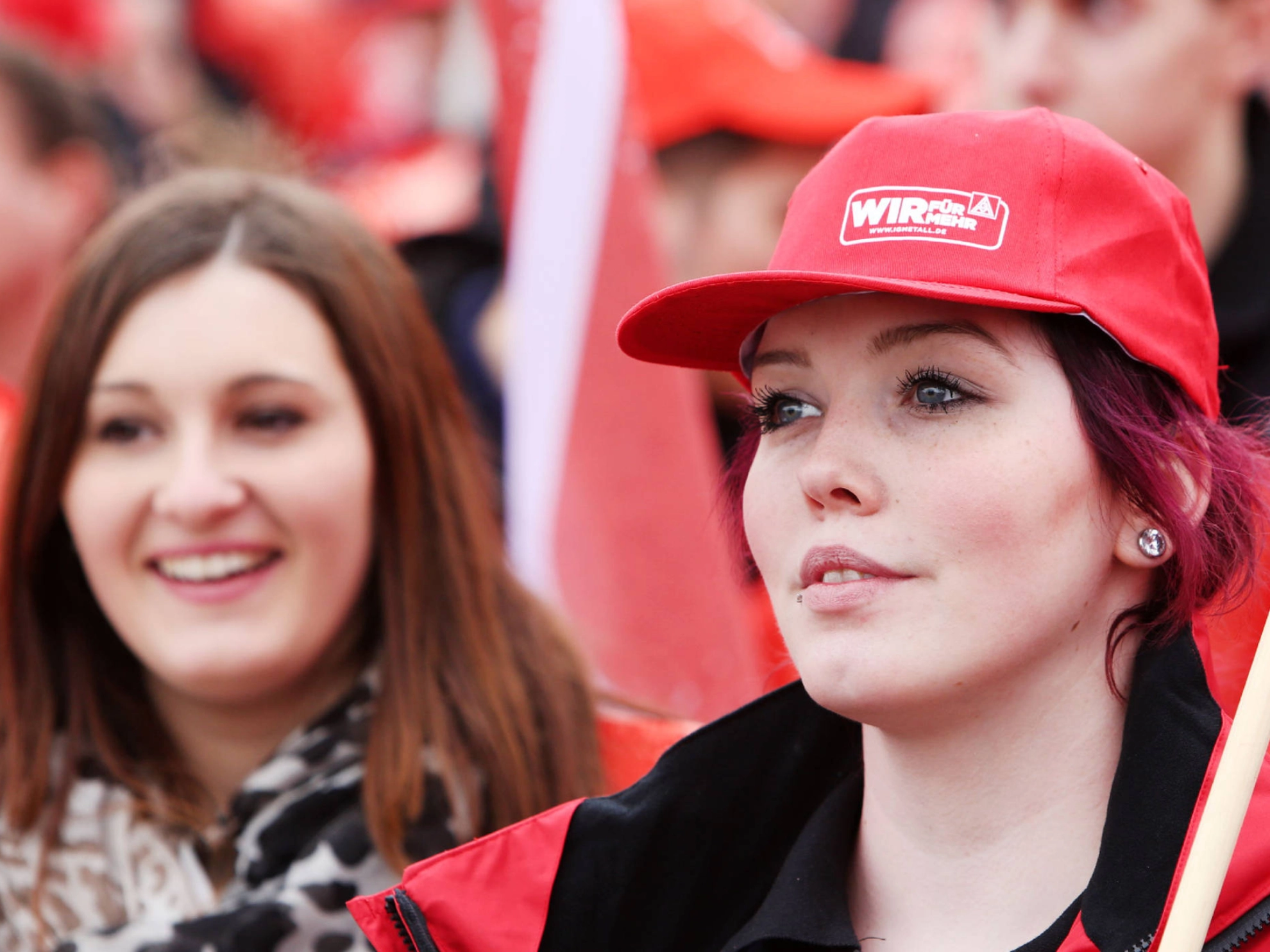 Zwei lächelnde Frauen, Eine trägt eine IG Metall-Kappe und eine Fahne über die Schulter.